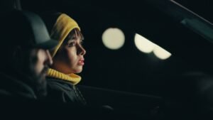 A woman in a yellow hood beside a man in a car at night in Passengers (2024).